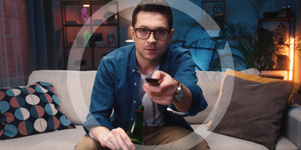 A man in glasses watching television choosing channel while sitting on sofa and drinking beer from bottle.
