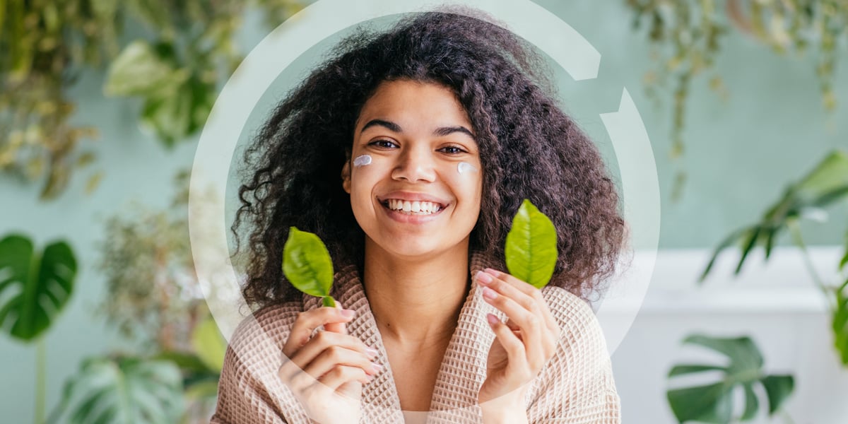 A woman holding 2 leaves, smiling, with skincare product on her face.